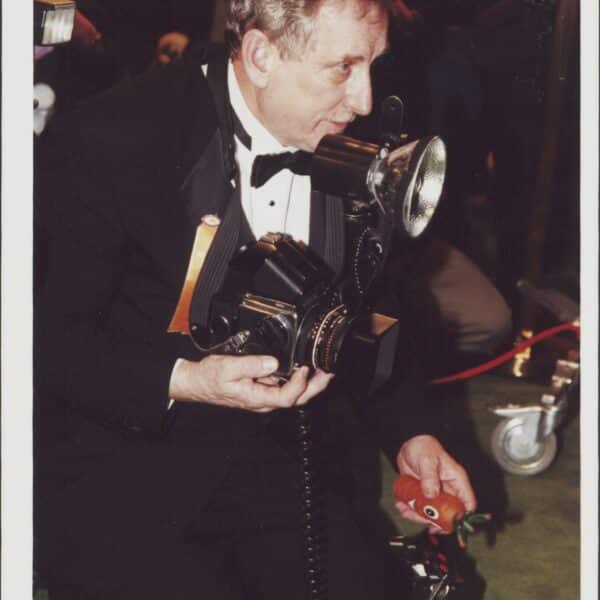 An older man in a tuxedo kneels on green carpet holding a vintage camera with a flash, surrounded by photography equipment.