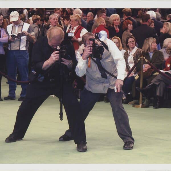 Two photographers with cameras crouch and take pictures at an indoor event, with a seated and standing audience in the background.