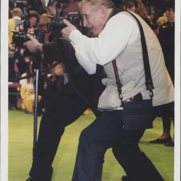 Two photographers with cameras and flash attachments crouch and take pictures at an indoor event, with several people visible in the background.