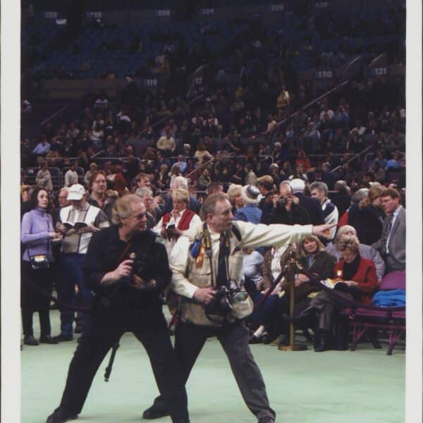 Two men stand in front of a crowd at an indoor event; one points ahead while the other stands close beside him, both holding cameras.