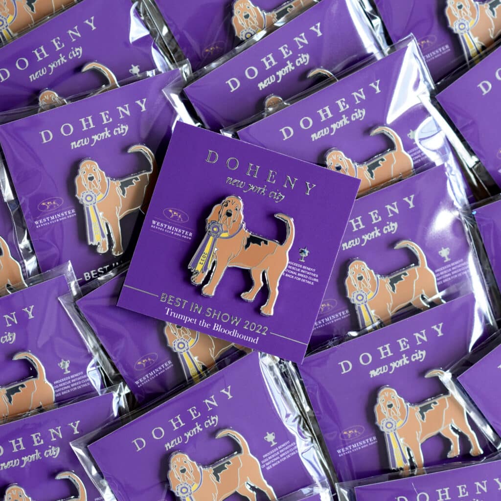 Multiple enamel pins featuring a bloodhound dog with a blue ribbon are displayed on purple cards labeled "DOHENY new york city" and "Best In Show! 2022 Trumpet the Bloodhound.