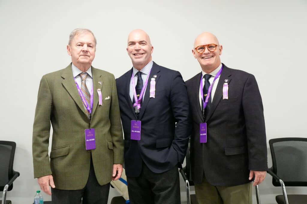 Three men in suits and purple lanyards stand together indoors in front of a white wall, two smiling and one with a neutral expression, at an event reminiscent of the professionalism seen at the Westminster Kennel Club.
