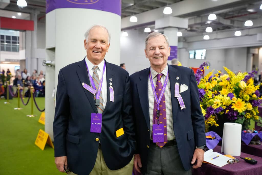 Two older men wearing blazers, ties, and purple all-access badges stand indoors near a display of yellow and purple flowers at a Westminster Kennel Club event.