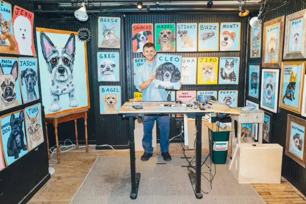 An artist stands at a desk in a studio filled with colorful dog portraits, holding up a painting of a black and white dog labeled "Cleo".