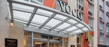 The entrance of a Voco hotel, featuring a glass canopy, modern exterior, and black and gold Voco Hotels signage above and beside the door.
