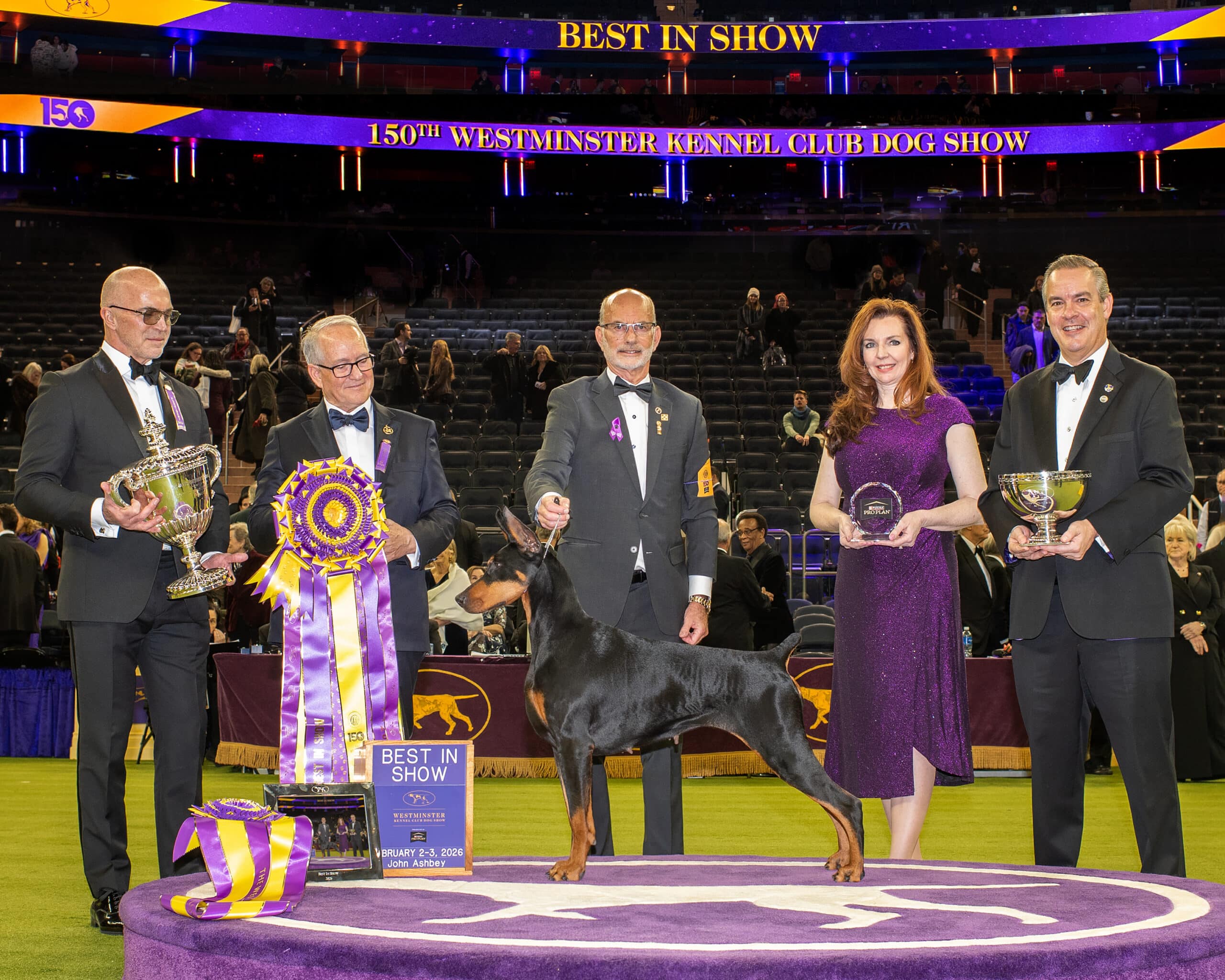 A group of five people stands with a Doberman Pinscher and trophies under a "Best in Show" sign, celebrating the Westminster Kennel Club Best in Show Winner 2025 at the 150th anniversary event.