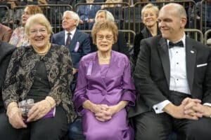 Three people in formal attire sit in the front row at an event, with more attendees seated behind them. The woman in the middle wears a purple dress.