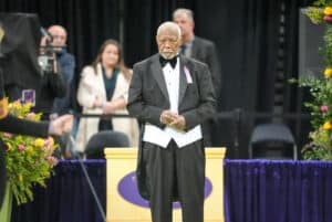 An older man in a tuxedo stands on a stage with flowers nearby, while people watch in the background.