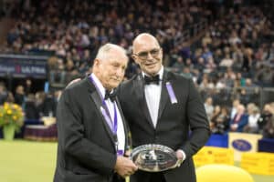 Two men in tuxedos stand on a green floor; one holds a glass award. A crowd is seated in the background at an indoor event.