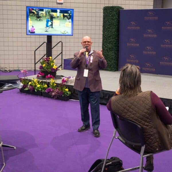 A man speaks into a microphone in front of a small seated audience, with a TV screen and flowers on a purple-carpeted stage behind him.