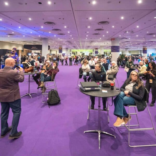 A speaker addresses an audience seated at round tables in a large convention hall with purple carpeting.