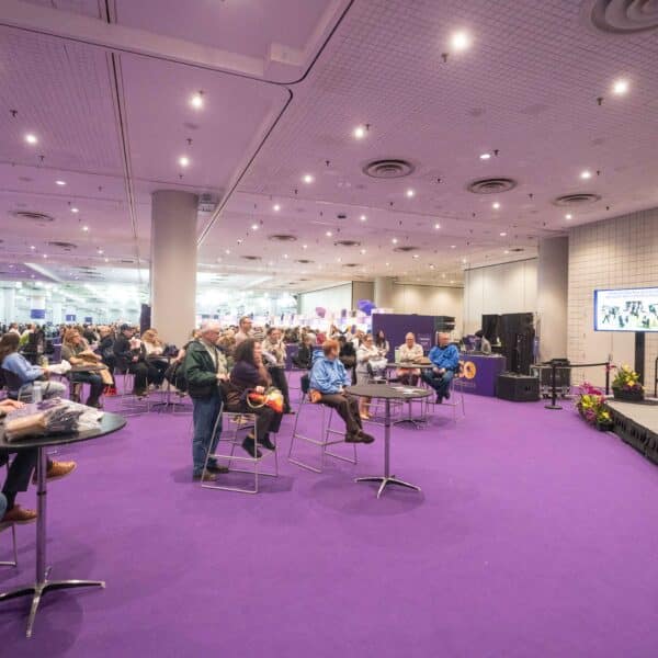 A group of people sit and listen to a speaker on a stage in a large room with purple carpet and overhead lights.