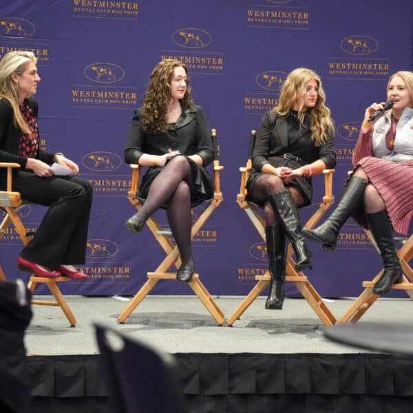 Four women sit on chairs on a stage, with one speaking into a microphone, in front of a Westminster Kennel Club Dog Show backdrop.