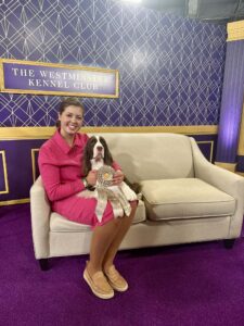 A woman in a pink outfit sits on a beige sofa holding a ribbon with a dog beside her at the Westminster Kennel Club event.