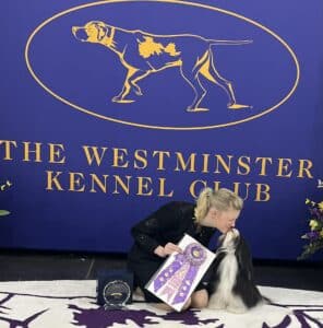 A woman kneels and kisses a dog, holding a large ribbon, with "The Westminster Kennel Club" logo displayed on a purple background behind them.