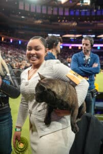 A woman in a white outfit holds a French Bulldog and a green ribbon at an indoor event with a large crowd and media personnel in the background.