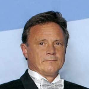 A middle-aged man with short brown hair wearing a tuxedo and bow tie stands in front of a blue background.