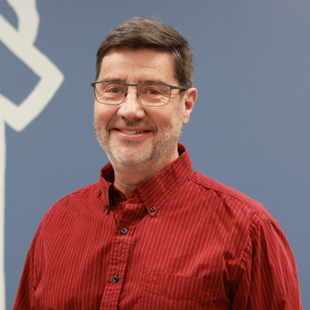 A man with short brown hair, glasses, and a beard—named Veterinarian of the Year—is wearing a red button-up shirt and standing in front of a blue wall with a white abstract design.
