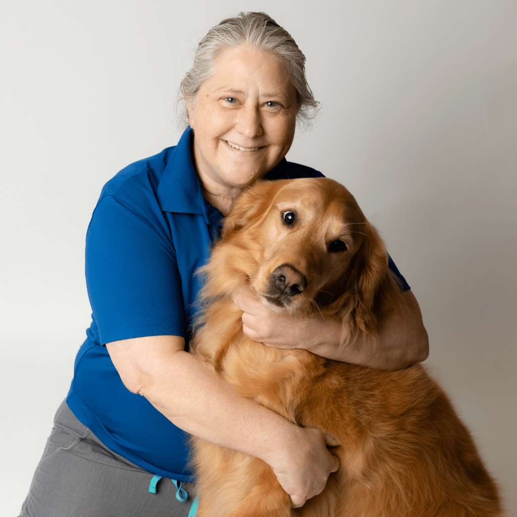 An older woman in a blue shirt, honored as Veterinarian of the Year, smiles while hugging a golden retriever against a plain light background.