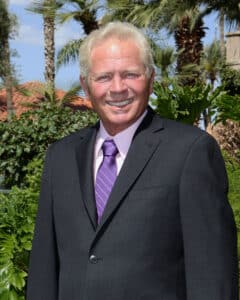 A man in a dark suit and purple tie stands outdoors in front of palm trees and greenery, smiling at the camera.
