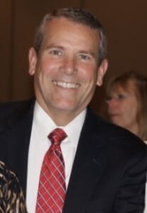 A man in a dark suit, white shirt, and red striped tie smiling at the camera, with a blurred woman in the background.