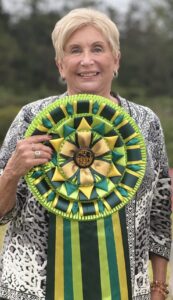 Two people pose outdoors; one holds a large green and yellow award ribbon, the other holds a white fluffy dog.