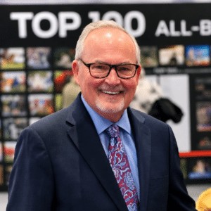 Smiling man in a suit and tie stands indoors with a "TOP 100 ALL-" display and photos visible in the background.