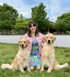 A woman in a colorful dress and pink jacket kneels on grass, smiling, with a golden retriever on each side. Trees and a building are in the background.