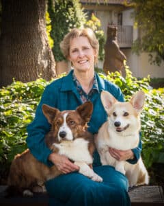 A woman in a teal jacket sits outdoors on a bench, smiling and holding two Corgi dogs, one brown and one light-colored, with greenery and a building in the background.