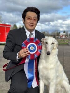 A man in a suit holds a large, multicolored award ribbon next to a white dog outdoors on a cloudy day.