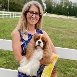 A woman with long gray hair and glasses smiles while holding a Cavalier King Charles Spaniel in front of a white fence and grassy field.