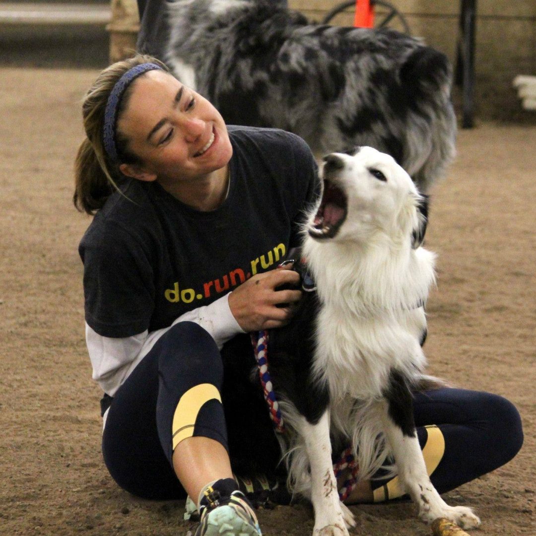 A person sits on the ground indoors with a black and white dog yawning beside them, embodying the kind of quiet, comforting moment you might find captured by the Veterinarian of the Year.